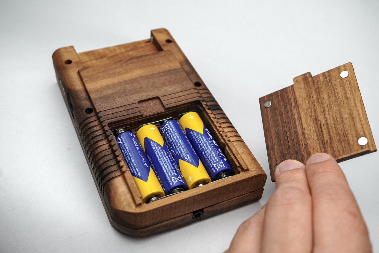 Photo of the backside of the wooden Game Boy. The battery compartment is open with batteries inside and a hand is holding its cover next to it. Four small magnets are clearly visible in the back of that cover.
