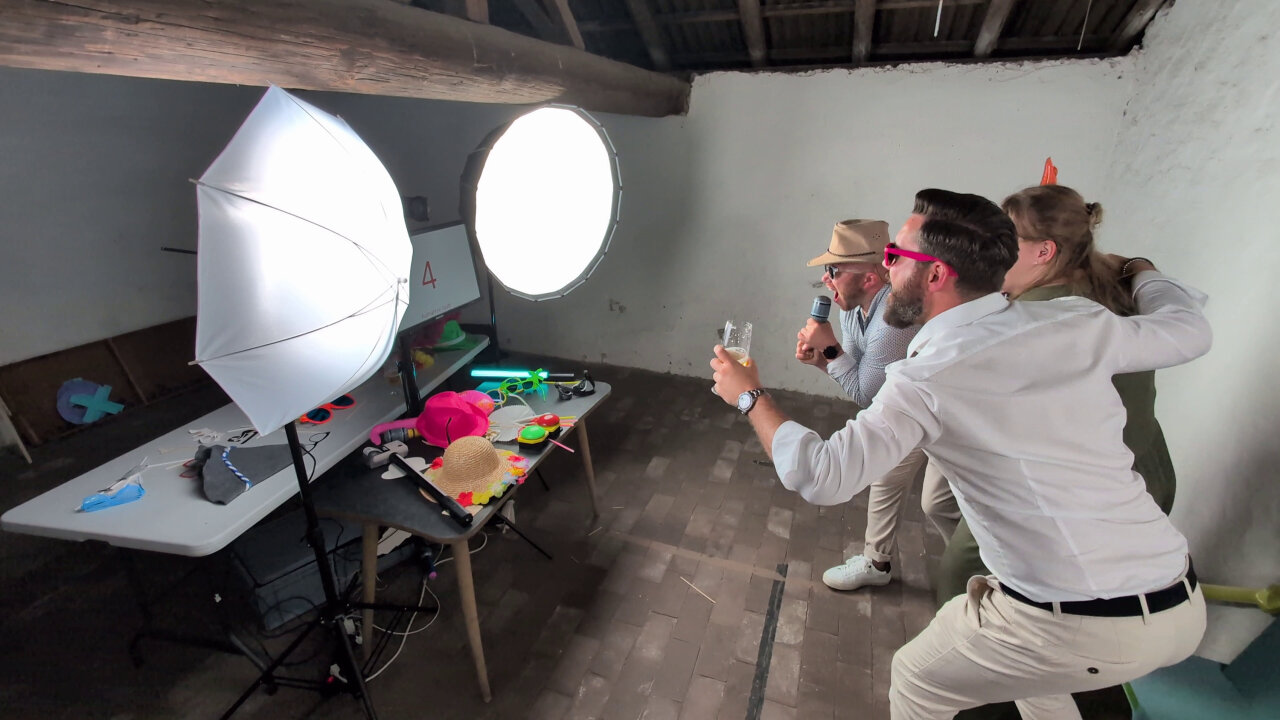 Wide angle shot of a small room. On the left there is a table with colorful disguises like hats and oversized sunglasses. On the table are also a red and green push button and a Game Boy Printer. Above the table are two large video lights an a screen inbetween those lights with a camera barely visible above the screen. On the right, in front of the camera, are three party guests with diguises singing, linking arms and looking at the camera.