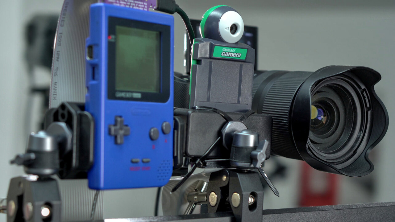 Detail shot of the camera assembly at the top of the photo booth. From left to right it shows a Game Boy Pocket, a Game Boy Camera in a mount and a Sony a6400 from which mostly a big Tamron lens is visible.