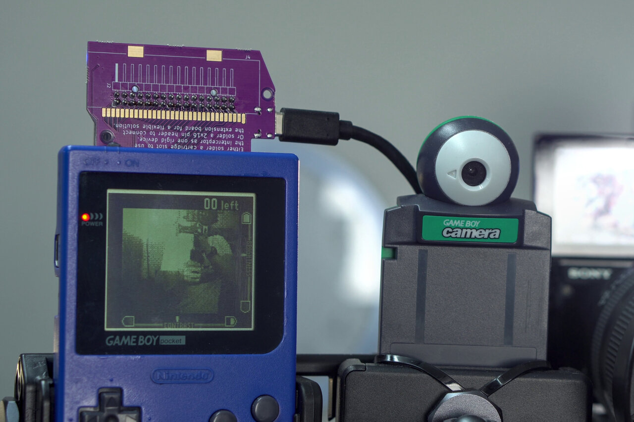 Detail shot of the Game Boy and the Game Boy Camera above the photo booth screen. The Game Boy on the left is a blue Game Boy Pocket with a purple PCB sticking out from its top. A USB-C cable is attached to the side of the PCB. On the right is a green Game Boy Camera, held to a mount with zip ties.