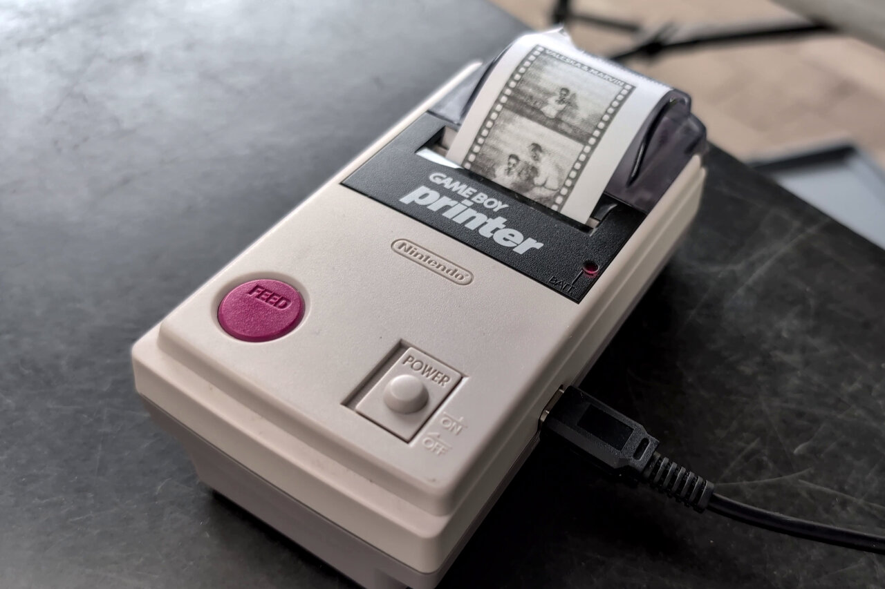 Close-up of the Game Boy Printer on the table at the wedding reception. A half-printed strip of paper is sticking out from its top, showing two stills from a video recorded by guests.