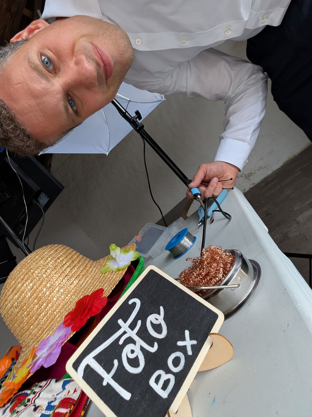 Photo of me in a white shirt holding a soldering iron next to a table with a sign Foto Box (in German) on it.