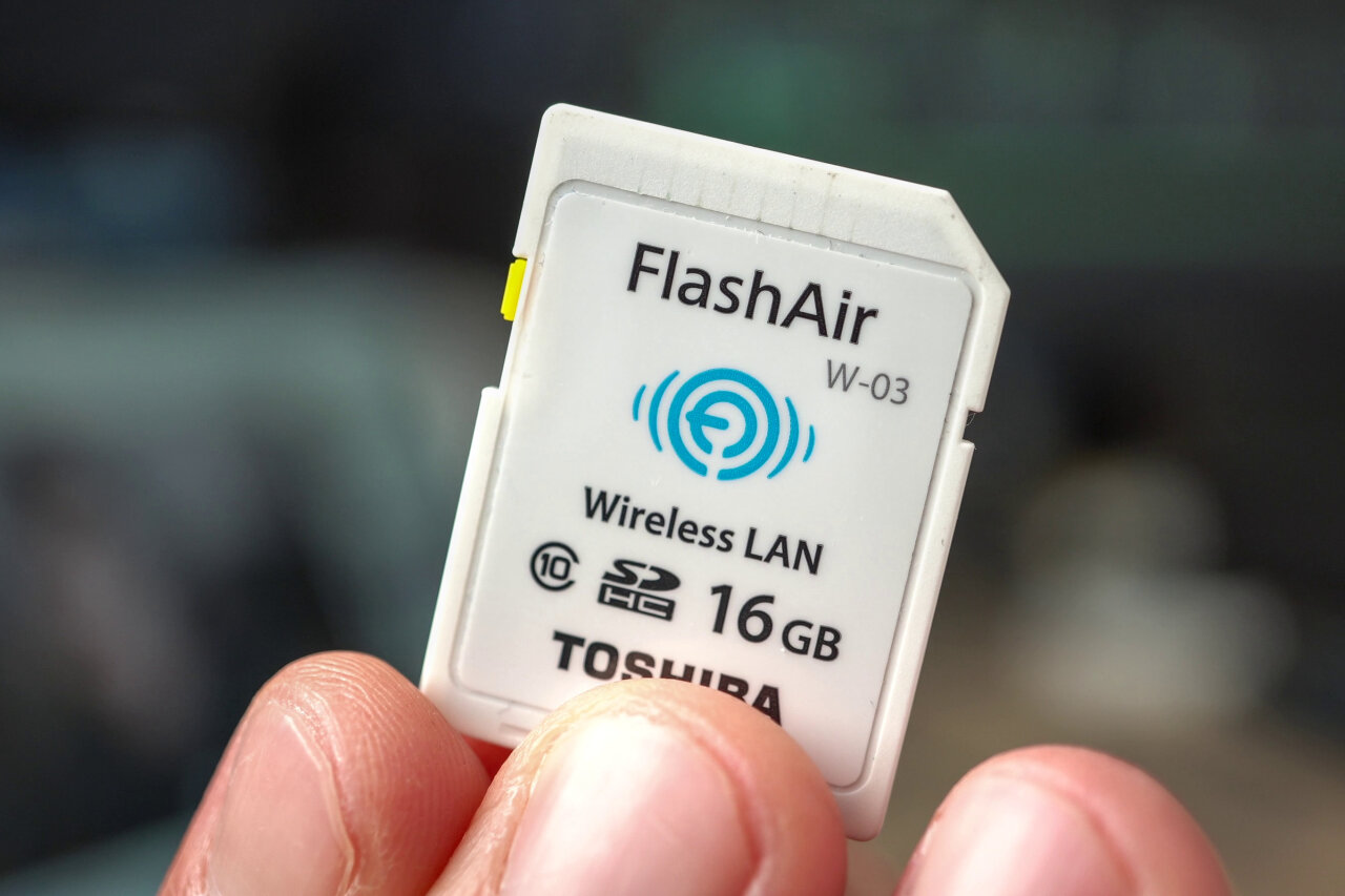 Photo of fingers holding a white SD card. Its label reads FlashAir W-03, Wireless LAN 16GB Toshiba.